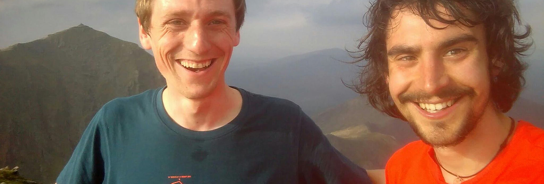 Two men are smiling at the camera, standing upon a mountain-top.