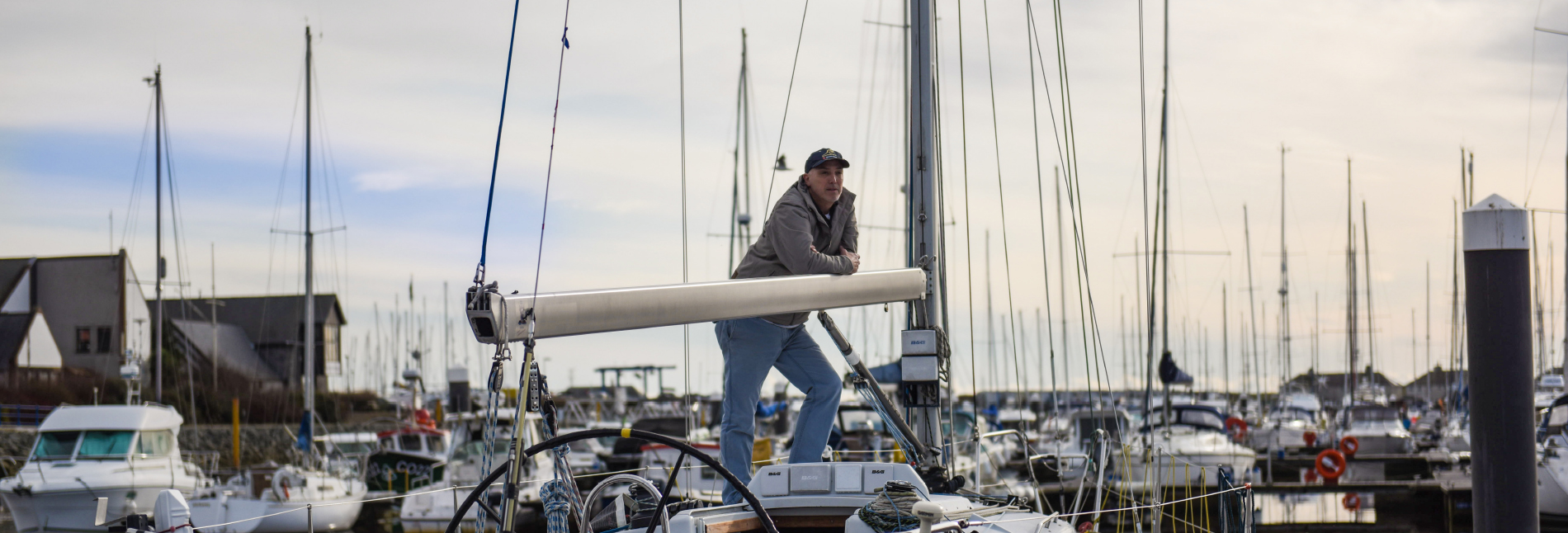 A man on a yacht looks to the distance. In the background is a harbour full of boats.